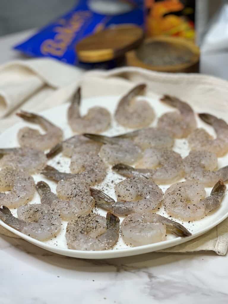 Raw, peeled and deveined shrimp with the tail on, seasoned with salt and pepper and laying on a white platter.