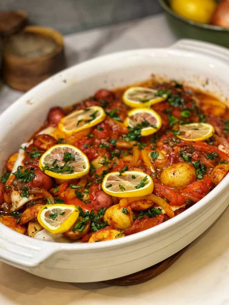 A casserole dish of oven roasted cod topped with thin slices of lemon and garnished with fresh chopped parsley.