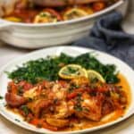 Oven roasted cod covered in a Mediterranean-style tomato sauce sitting on white plate with a side of sautéed spinach.