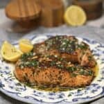A white plate with a blue patterned holding two seared salmon filets topped with a lemon butter garlic sauce.