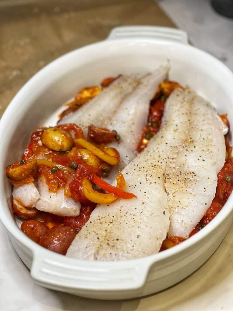 Two seasoned cod filets laying in a casserole dish that is lined with a tomato/vegetable sauce.