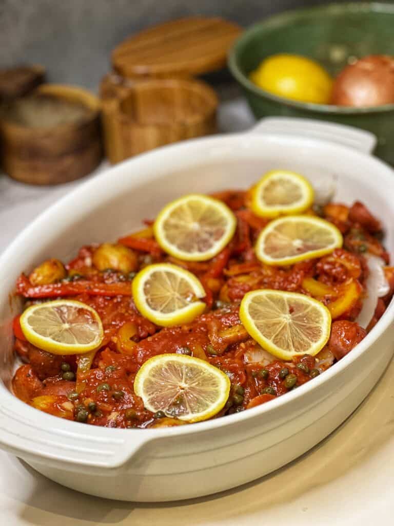 A casserole dish of cod covered in a tomato sauce topped with thinly sliced lemons.
