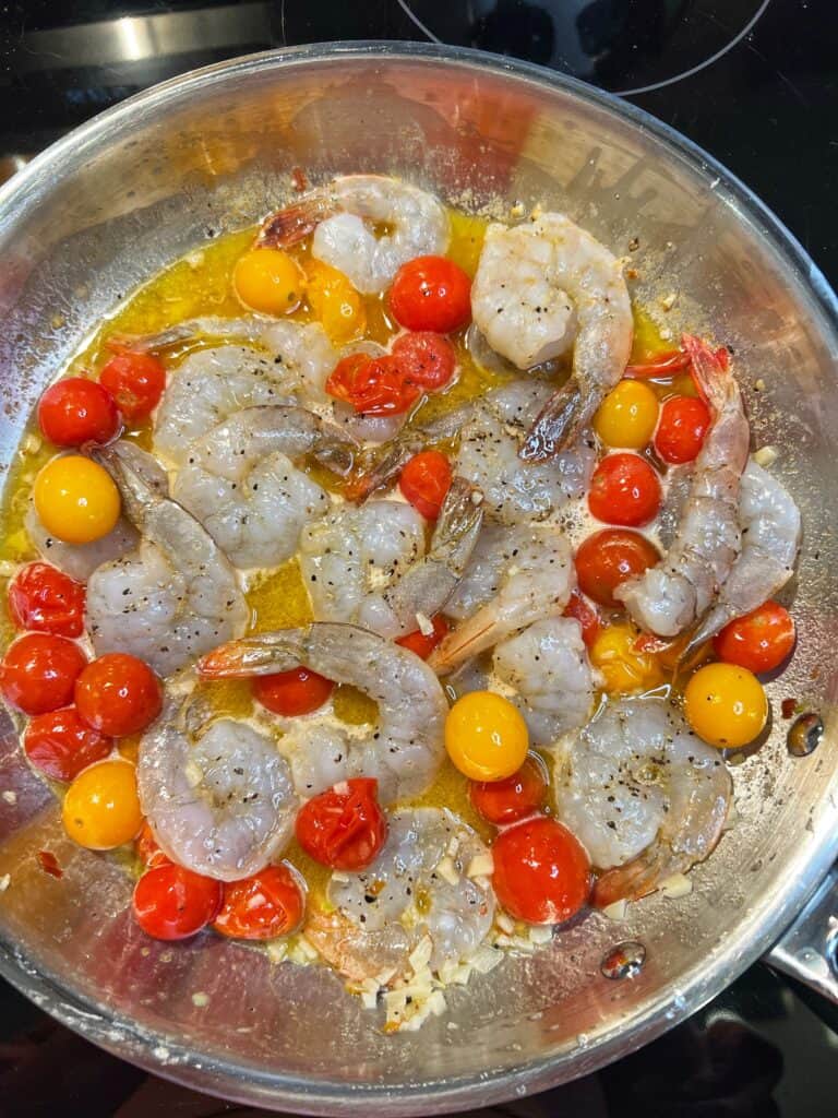 Raw shrimp added to a stainless steel pan with bursting cherry tomatoes.
