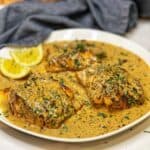 A white platter of three blackened cod filets covered with a creamy pan sauce and garnished with fresh chopped parsley.