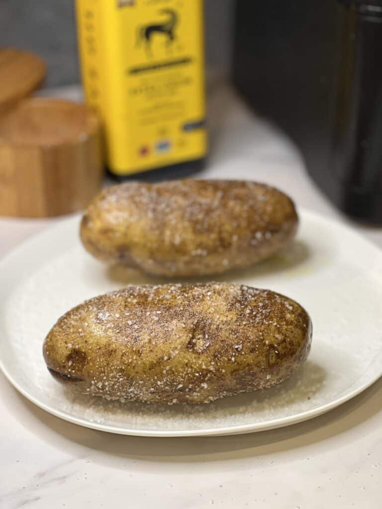 Two russet potatoes sitting on a white plate that have been rubbed with olive oil and coated in kosher salt.