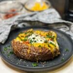 An air fryer whole "baked" potato sitting on a charcoal-colored plate loaded with cheese, bacon, sour cream and fresh parsley.
