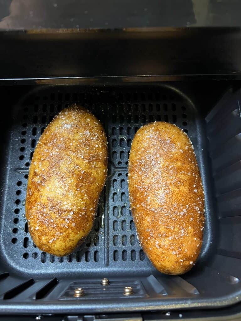 Two russet potatoes that have been coated with olive oil and salt sitting in an air fryer basket.