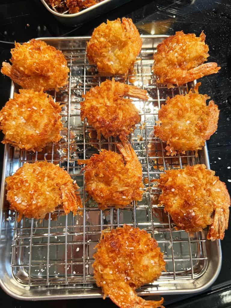 Ten crispy fried coconut shrimp sprinkled with salt and laying on a baking rack fitted to a small sheet pan.