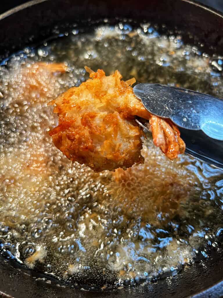 Holding a piece of crispy fried coconut shrimp over a pot of oil using stainless steel tongs.