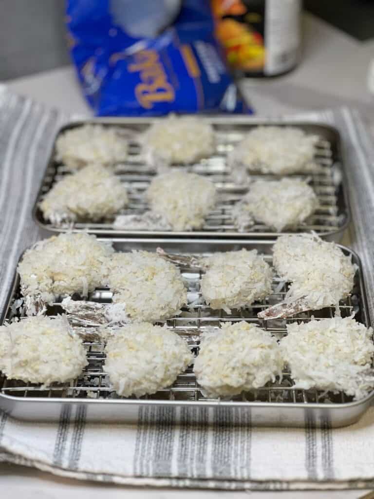 Shrimp coated in a shredded coconut/panko mixture sitting on two small baking pans fitted with racks.