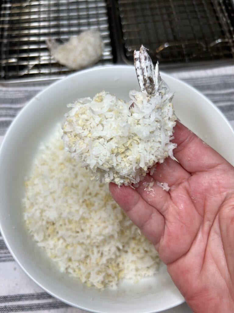 Me holding a piece of tail-on shrimp that has been coated in a shredded coconut and panko mixture.