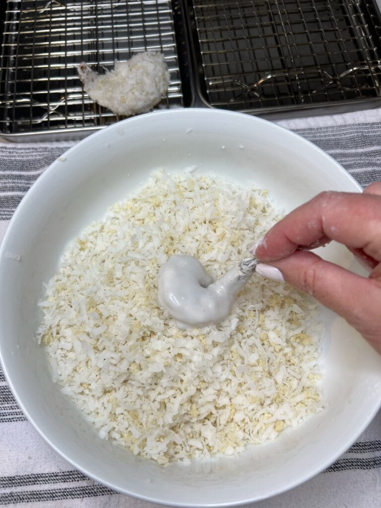 A coconut cream coated shrimp being placed in a white bowl of shredded coconut and panko.