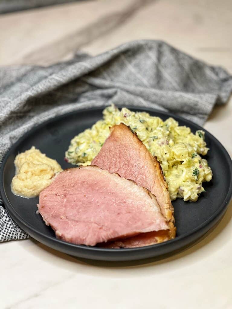 Two slices of ham on a charcoal colored plate with a side of potato salad and a spoonful of horseradish.