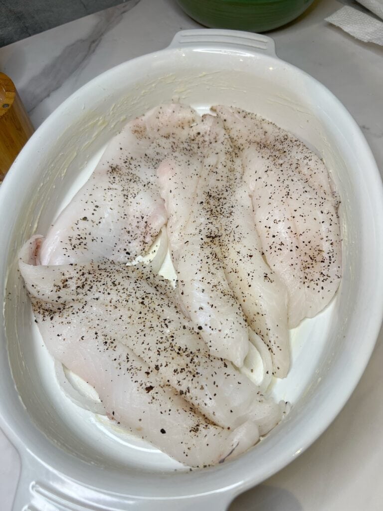 Four seasoned orange roughy filets sitting on top of sliced white onions in an oven-safe casserole dish.