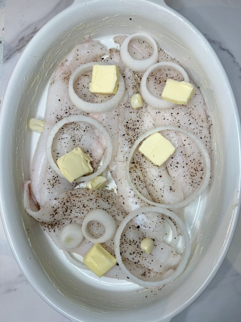 Five pats of butter, sliced white onions and garlic cloves added to the top of seasoned orange roughy filets that are sitting in an oven-safe casserole dish.
