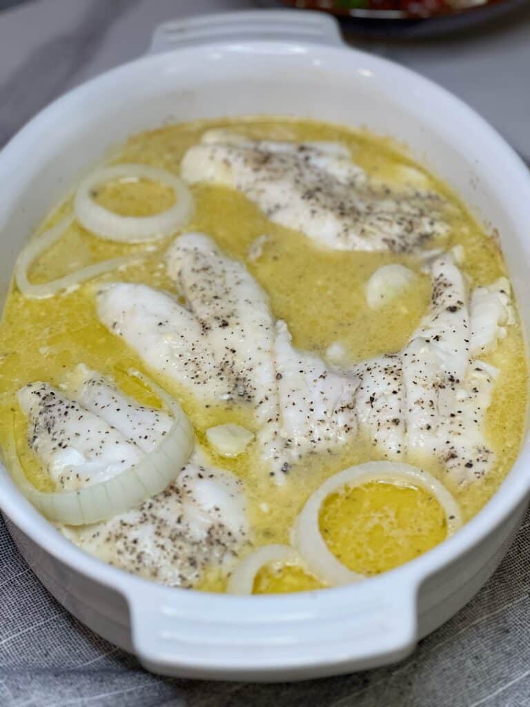 A white casserole dish of oven poached orange roughy. The poaching liquid is yellow in color from the combination of butter and orange juice.