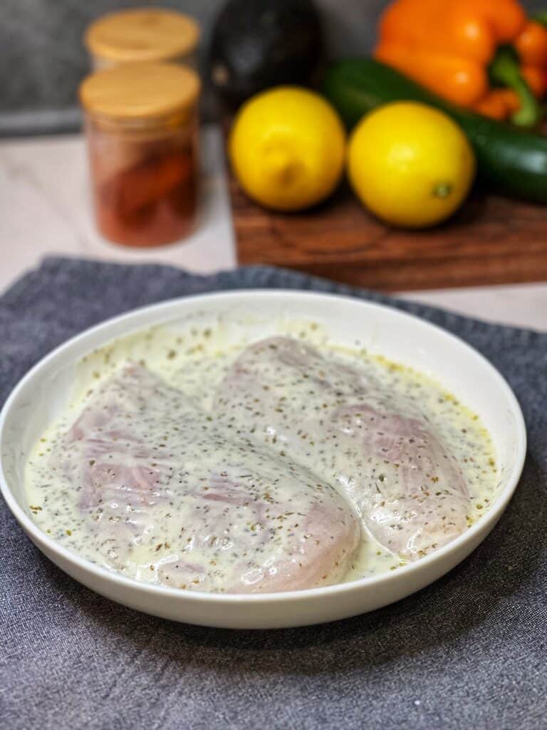 Both sides of two raw chicken breasts in a white bowl covered in a Greek-style marinade for Greek Chicken Rice Bowls.