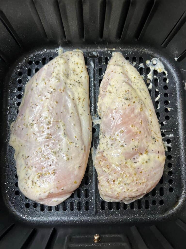 Two raw chicken breasts coated in a Greek marinade sitting in an air fryer basket.