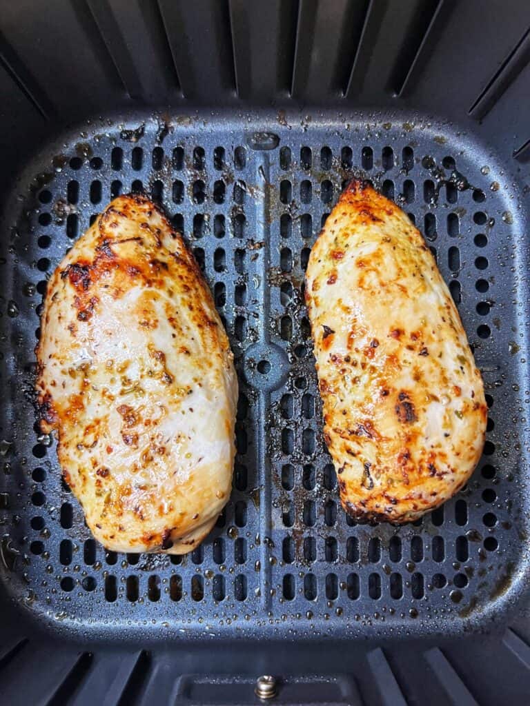 Two air fried chicken breasts sitting in an air fryer baskets for Greek Chicken Rice Bowls.