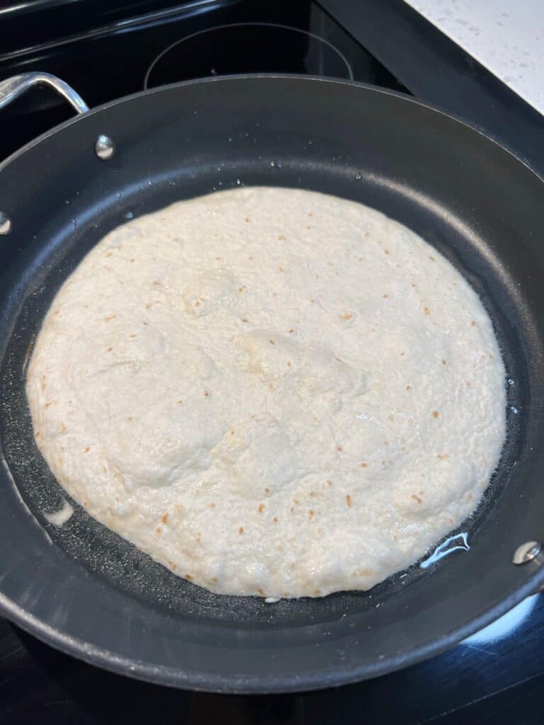 A burrito-sized flour tortilla frying in a non-stick pan with oil.