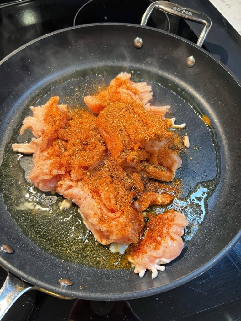 Ground chicken sprinkled with taco seasoning cooking in a non-stick pan with heated olive oil.
