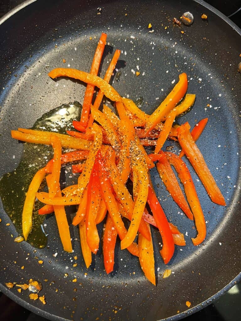 Sliced red and orange bell peppers sautéing in olive oil in a non-stick pan.