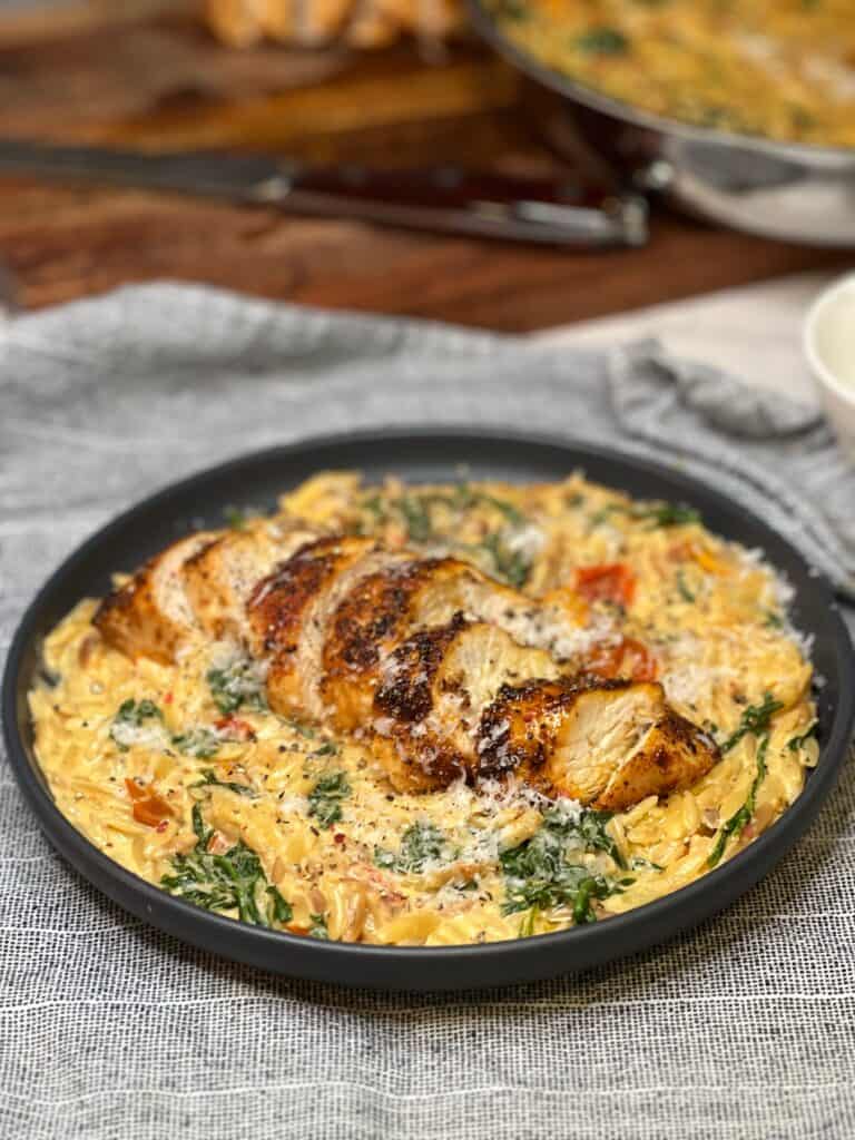 A cut up chicken breast laying on top of a bed of creamy tomato orzo pasta on a charcoal-colored plate. The dish is garnished with freshly grated Parmesan cheese.
