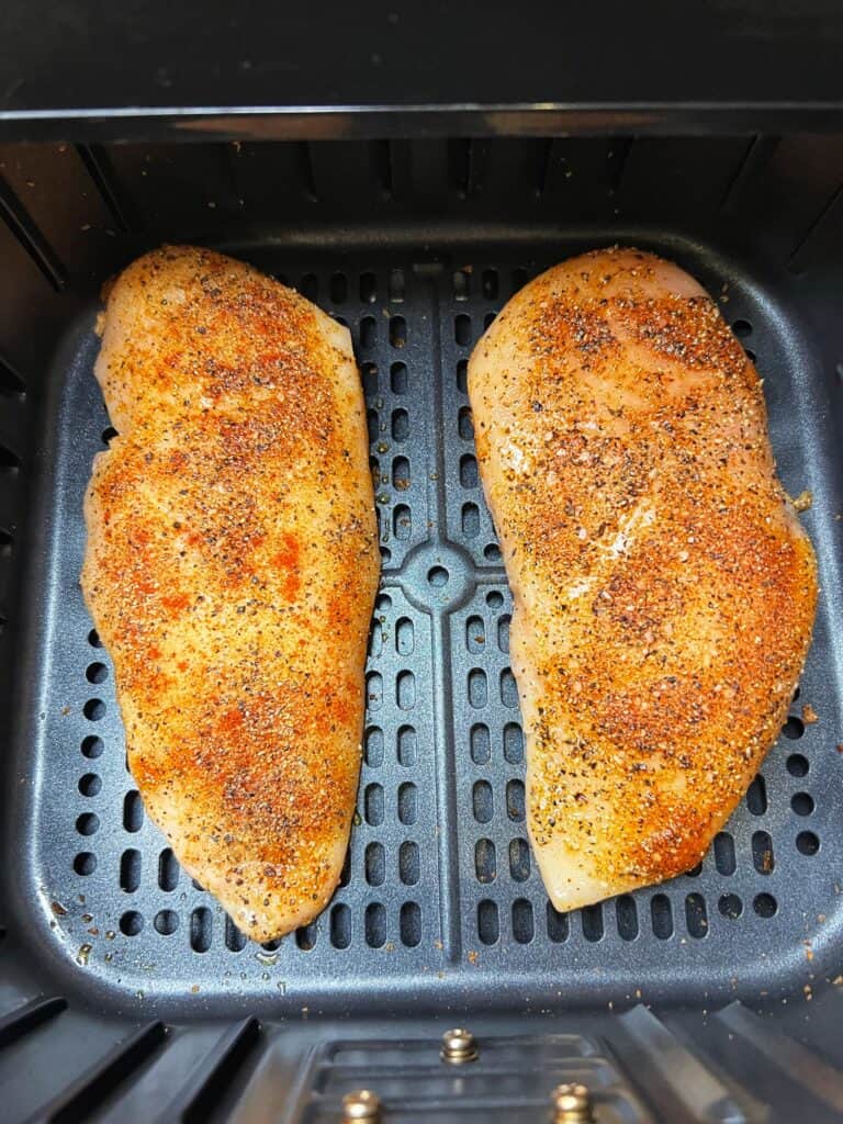 Two raw, seasoned boneless chicken breasts in the basket of an air fryer.
