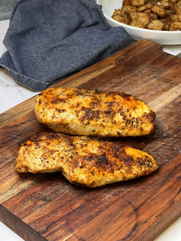 Two seasoned and fully-cooked, boneless chicken breasts made in the air fryer sitting on a wooden cutting board.