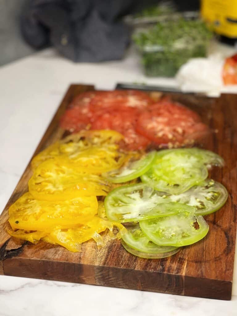 Thin slices of yellow, green and red heirloom tomatoes sitting on a wooden cutting board.