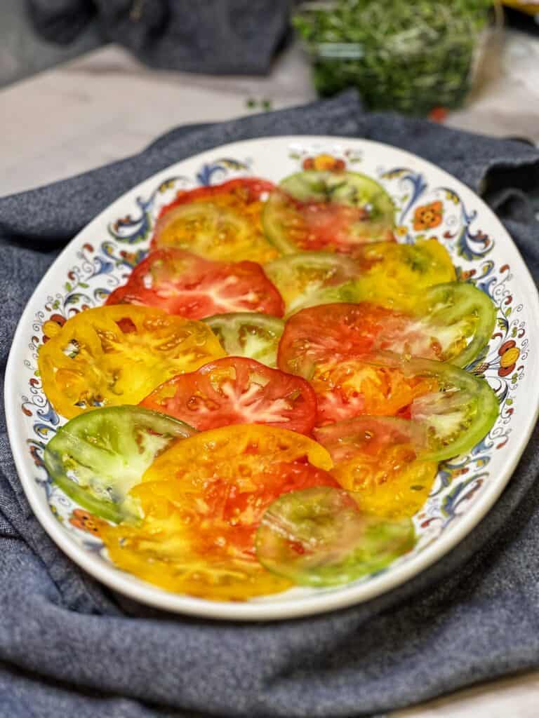 Thinly sliced heirloom tomatoes in a variety of colors arranged on a white platter with a blue decorative design on the edge.
