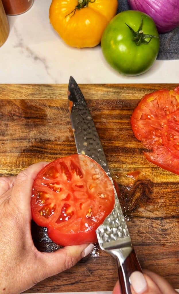 A red heirloom tomato sitting on a wooden board with the cut side up with a knife cutting a thin slice from the tomato.