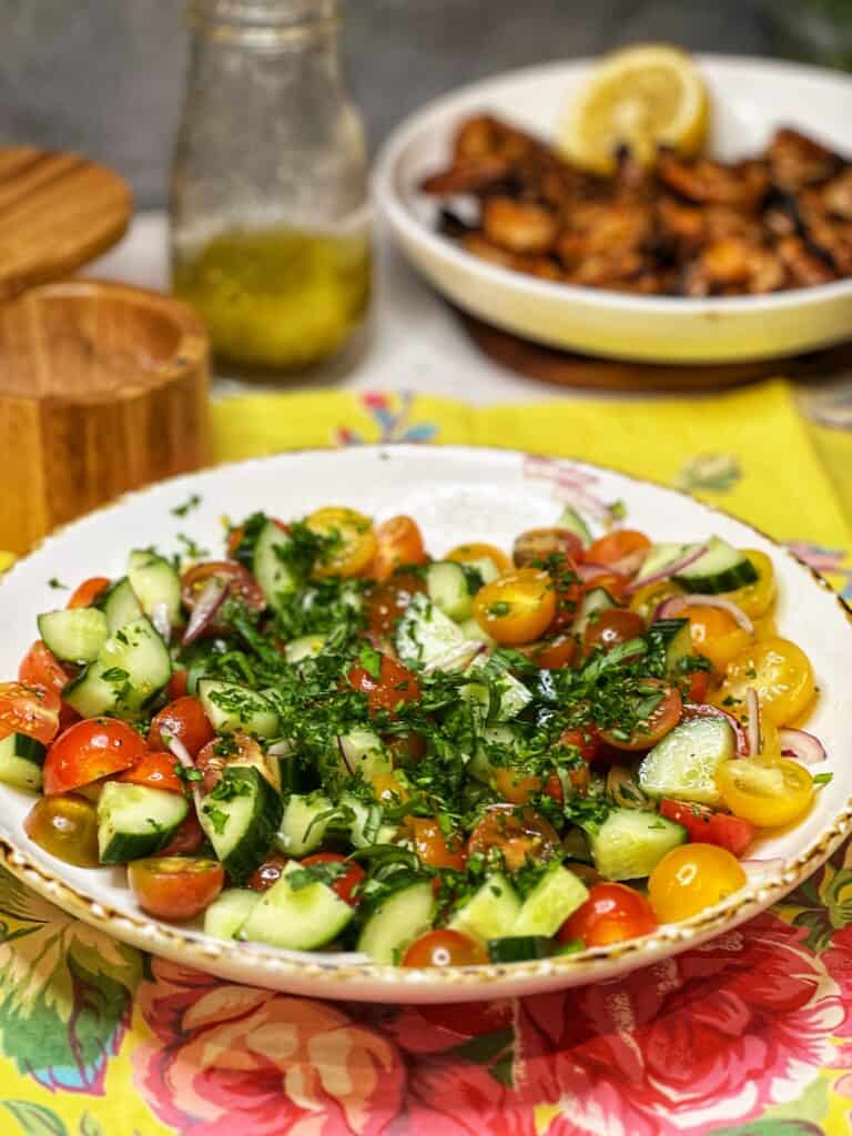 Halved cherry tomatoes, quartered cucumbers, red onion and fresh herbs in a white bowl for Healthy Grilled Shrimp Salad.
