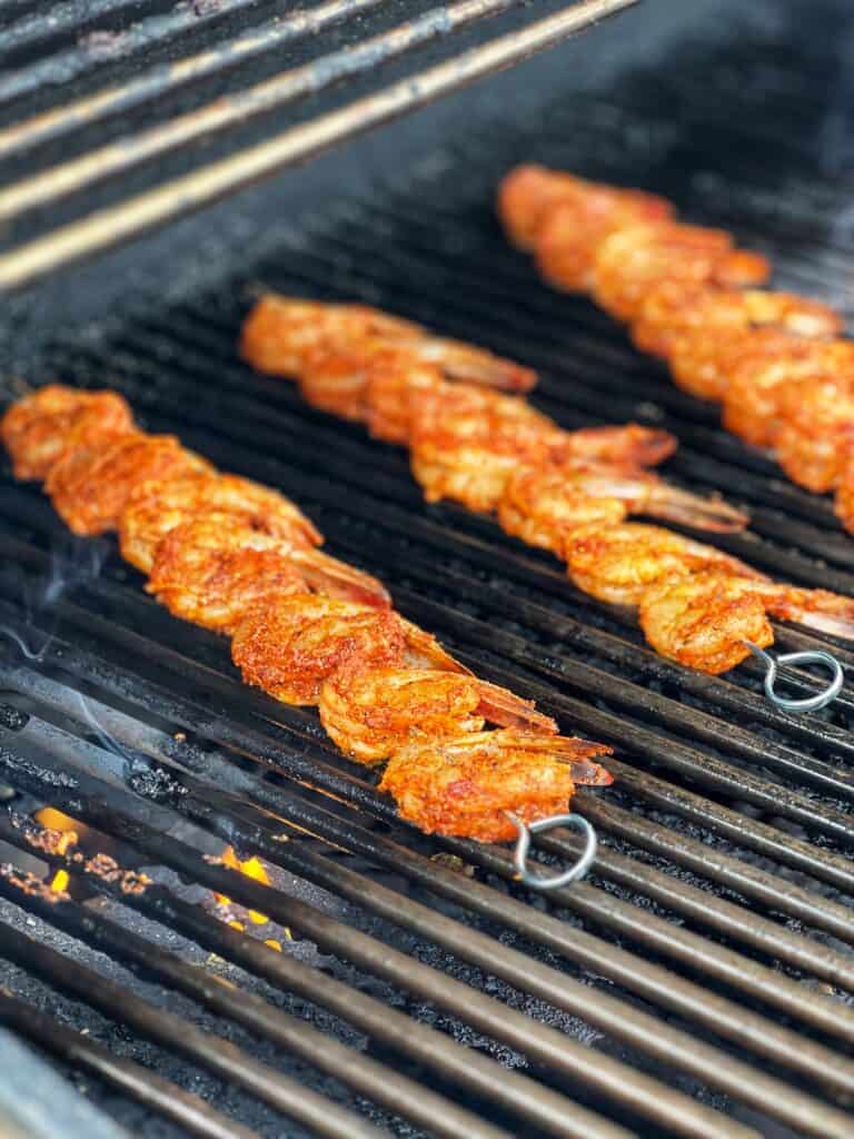 Seasoned shrimp on three metal skewers cooking on a grill. 