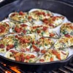 A cast iron pan sitting on a grill with twelve Rockefeller Style Oysters inside sitting on a bed of rock salt.