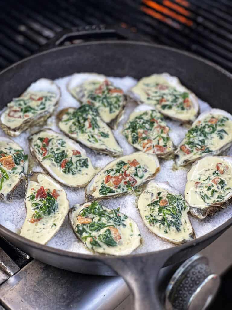 A spinach cream filling inside twelve raw oysters sitting on a bed of rock salt in a cast iron pan that is on a grill.