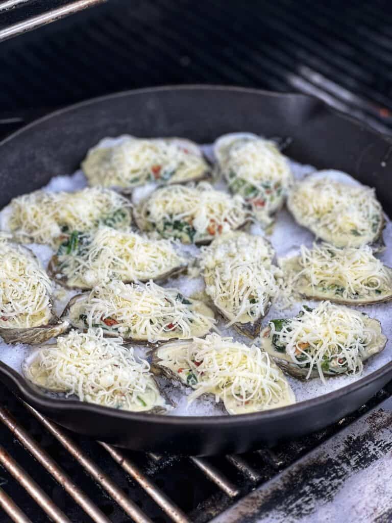 Cheese topped on raw oysters filled with a spinach cream sauce for Rockefeller Style Grilled Oysters. The oysters are sitting in a bed of rock salt in a cast iron pan on the grill.
