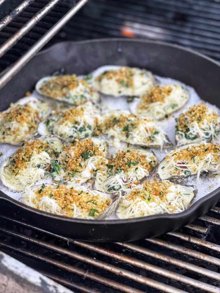 Twelve spinach cream filled oysters topped with cheese and toasty panko. The oysters are laying on a bed of rock salt in a cast iron pan on the grill.