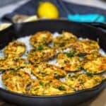 Twelve Creole Crab Baked Oysters sitting on a bed of rock salt in a cast iron pan. The oysters are filled and baked with a Creole mixture with crab. They have an orangish hue garnished with fresh parsley.