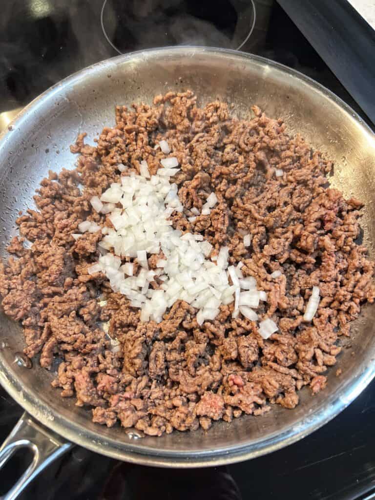 A stainless steel pan of browned ground beef with diced onions for Sloppy Joe Nachos.
