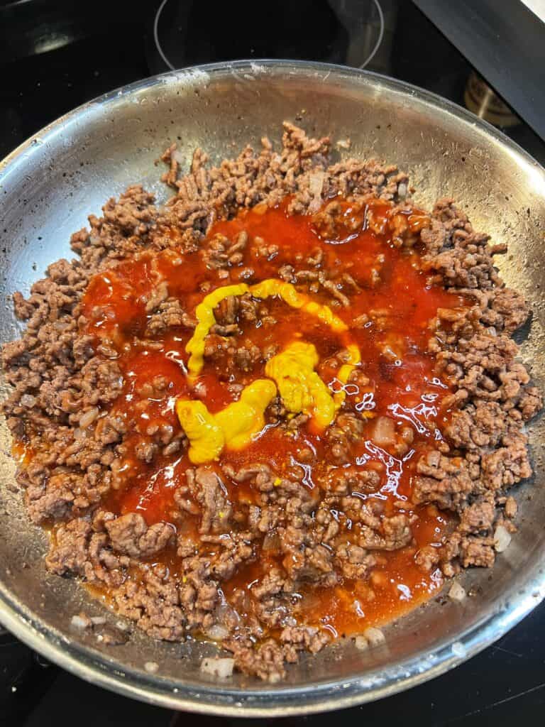 Ketchup and mustard added to a stainless steel pan of cooked ground beef.