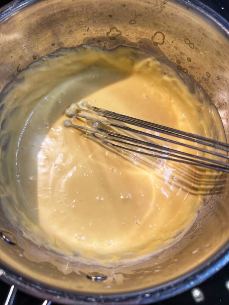 A saucepan of homemade cheese sauce with a wire whisk inside the pot.