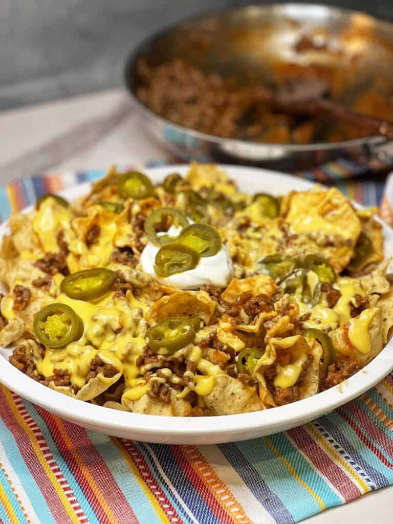 A large white bowl style platter filled with scoops tortilla chips covered in sloppy joes and homemade cheese sauce. All of that is dotted with jarred jalapeño slices and a dollop of sour cream.