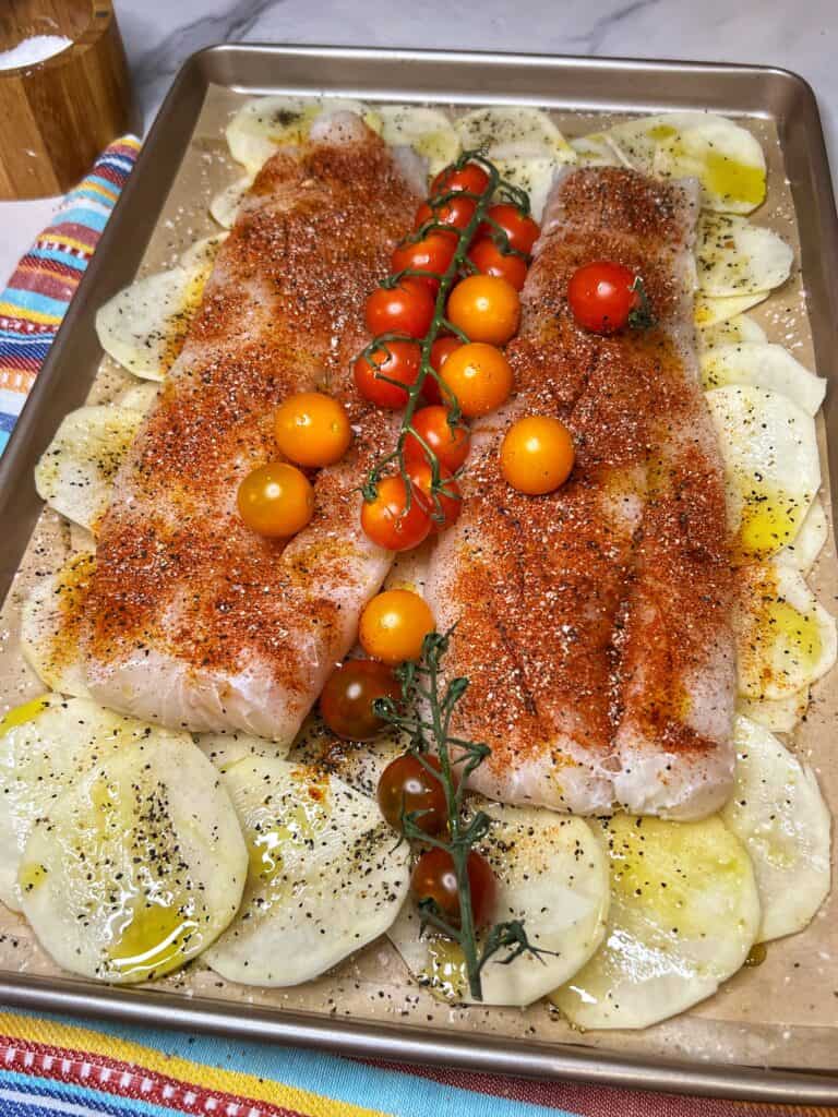 A large sheet pan layered with thinly sliced potatoes and topped with two large pieces of seasoned cod and cherry tomatoes ready to bake.