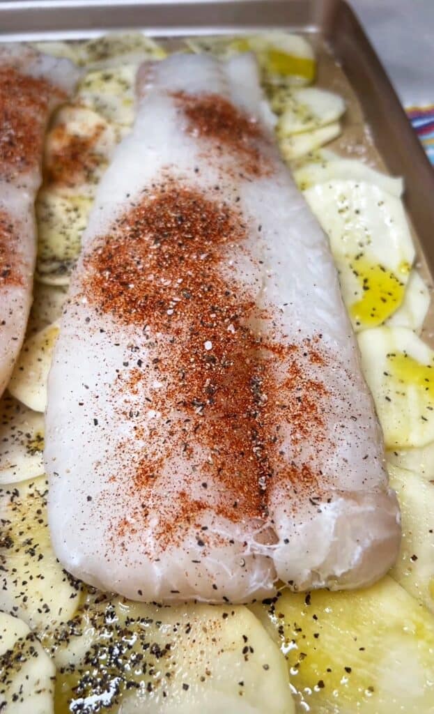A large piece of seasoned cod resting on top of a bed of thinly sliced potatoes in a baking sheet for Baked Cod with Brown Butter