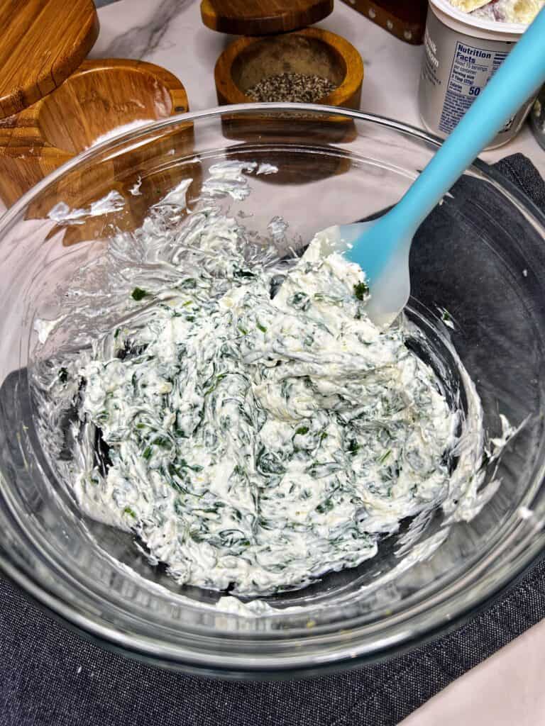 A creamy spinach mixture in a glass bowl with a blue spatula in the bowl.