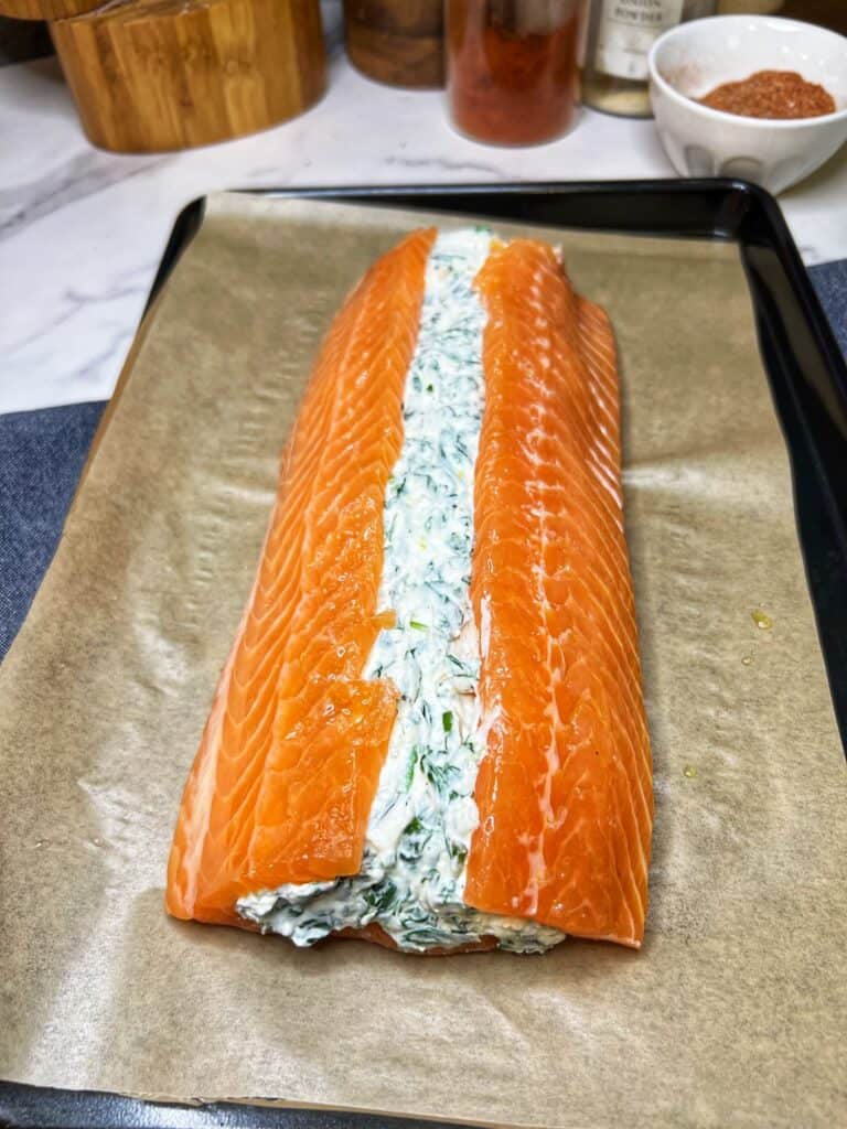 A filet of salmon sitting on a parchment-lined baking sheet. The filet is filled in the center with a creamy spinach mixture.