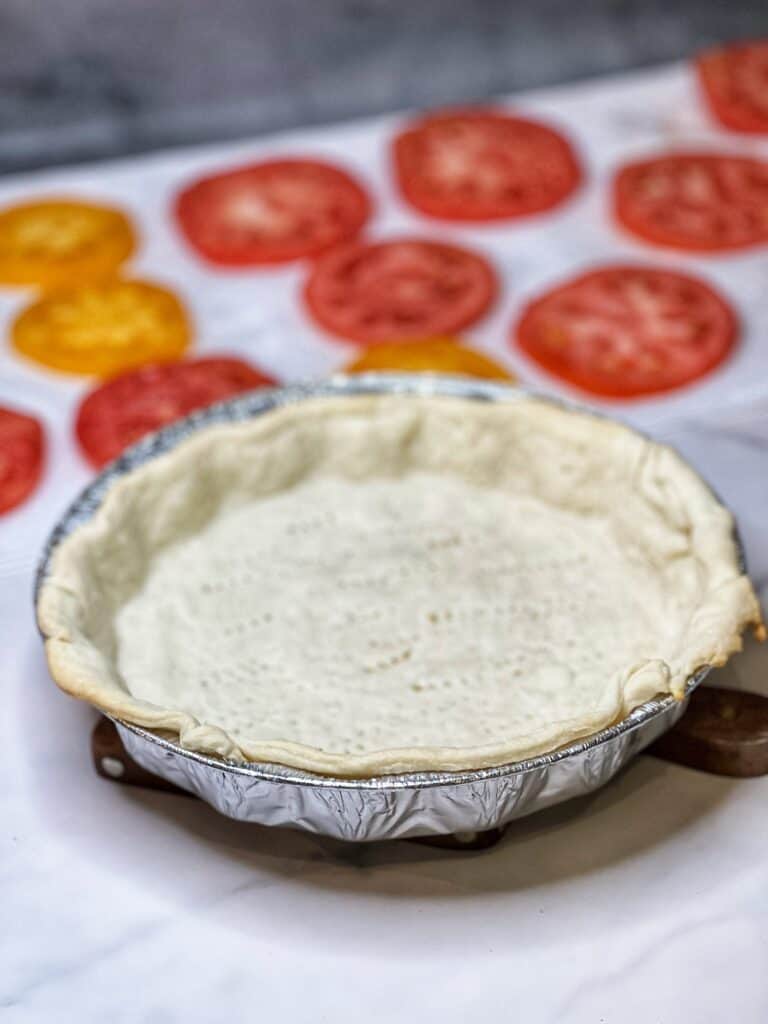 A par-baked pie crust sitting on a wooden board with sliced tomatoes in the background.