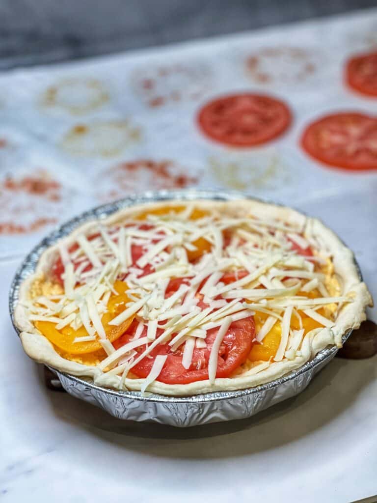 An heirloom tomato pie ready to be baked. The top layer contains slices of tomatoes sprinkled with white cheddar cheese.