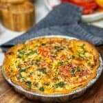 A Southern Heirloom Tomato Pie baked in an aluminum pie tin sitting on a wooden cutting board. The pie is garnished with fresh chopped basil.